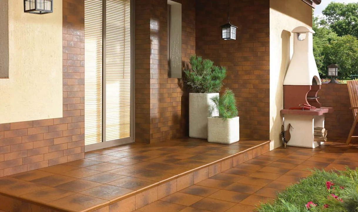 Taras z terakotowymi płytkami kamieniopodobnymi na podłodze i murkach oraz grillem z czerwonej cegły.