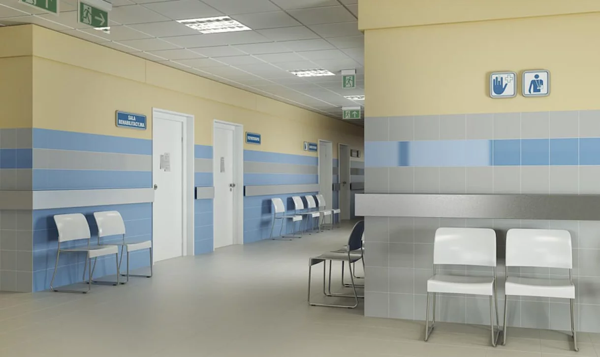 Korytarz szpitalny z poziomymi pasami niebiesko-szarych płytek kamieniopodobnych i białymi krzesłami.