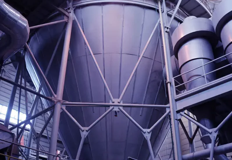 Wnętrze hali przemysłowej z dużym metalowym silosem i stalową konstrukcją wsporczą.