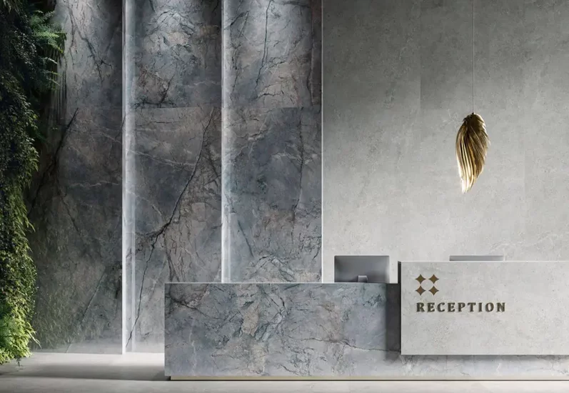 Recepcja z dużymi płytkami marmurowymi na ścianie i ladzie oraz złotą lampą i zielonymi roślinami.