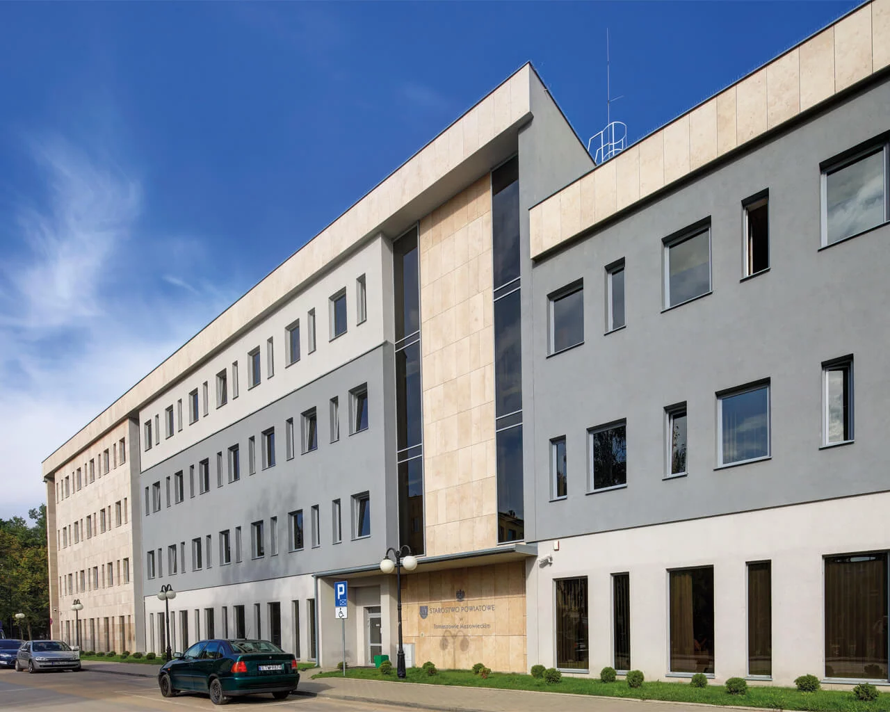 Elewacja nowoczesnego budynku publicznego z jasnobeżowymi płytkami kamieniopodobnymi i szarym tynkiem.